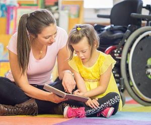 A little girl with cerebral palsy is sitting and listening to her caregiver read her a book at school.