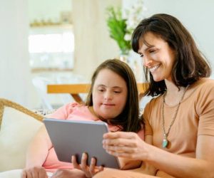 Smiling mid adult Hispanic woman and woman with Down Syndrome sitting side by side on family sofa and using digital tablet.